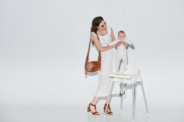 stylish woman in white dress and sunglasses carrying baby girl near high chair on grey