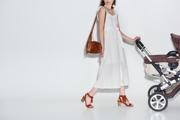 cropped shot of young woman in fashionable white dress walking with baby carriage on grey