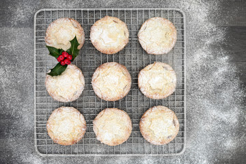 Freshly baked Christmas mince pies on a baking rack with holly and berry leaf sprig with icing sugar dusting on rustic wood background