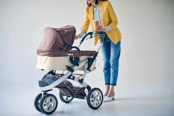 cropped shot of fashionable woman holding disposable coffee cup and standing with baby carriage on grey