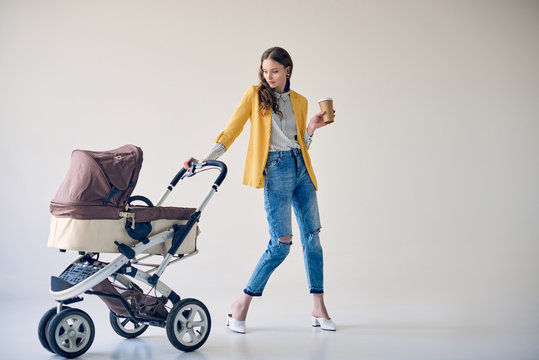 stylish young woman holding disposable coffee cup and looking at baby stroller on grey