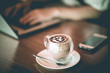 Hot Chocolate on glass  in coffee shop cafe with woman working with laptop in background,Selective Focus