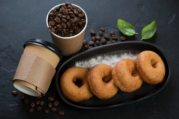 Cast-iron serving pan with donuts and takeaway carton coffee cups with coffee beans, studio shot on a black stone background