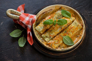 Baking pan with freshly cooked spinach and cheese cannelloni topped with green basil leaves, dark brown stone background