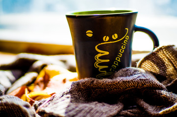 Fototapeta na wymiar A black cup with coffee near the window, with autumn yellow leaves and a knitted brown scarf on a wooden texture, on a sunny bright autumn day