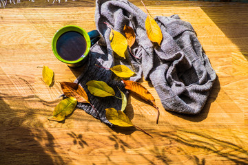 A black cup with coffee near the window, with autumn yellow leaves and a knitted brown scarf on a wooden texture, on a sunny bright autumn day
