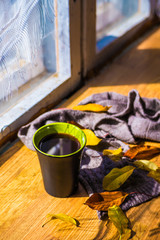A black cup with coffee near the window, with autumn yellow leaves and a knitted brown scarf on a wooden texture, on a sunny bright autumn day