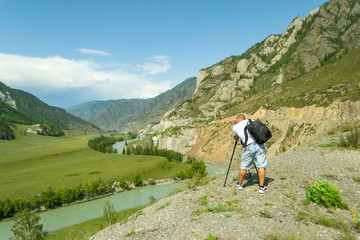 Naklejka premium Young man photographer travels through the Altai with the camera on the tripod takes a shot of the snow-covered mountains and the rocks with turquoise winding river Katun. Photo tour.