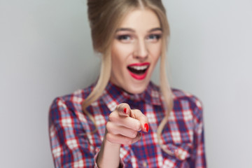 Amazed beautiful blonde girl with pink checkered shirt, collected hairstyle and makeup standing, looking at camera and with finger pointing and shocked. indoor studio shot, isolated on gray background