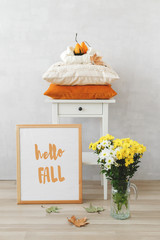 Autumn home interior decor. Bedside table, frame with text HELLO FALL, vase with white and yellow chrysanthemums flowers, orange and beige pillows, pumpkins, dry leaves. Cozy modern stylish room.