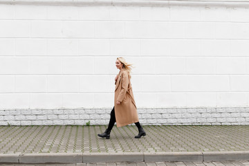 Beautiful young stylish blonde woman wearing long beige coat, black boots and black jeans walking through the city streets. Trendy casual outfit. Street fashion.