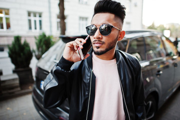 Stylish indian beard man at black leather jacket and sunglasses against business suv car. India model posed outdoor at streets of city and speaking on phone.