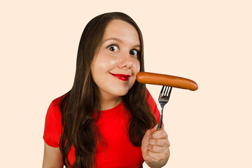 Young cute girl holding a sausage on a fork at her mouth, isolated on a light pink background