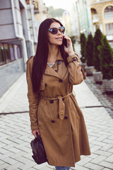 Nice to hear you! Beautiful young woman in trench looking away with smile and talking on mobile phone while standing outdoors