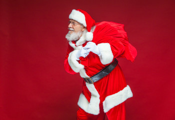 Christmas. Kind and tired Santa Claus in white gloves carries a red bag with gifts over his shoulder. Isolated on red background.