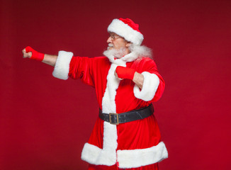 Christmas. Santa Claus with red bandages wound on his hands for boxing imitates kicks. Kickboxing, karate, boxing. Isolated on red background.