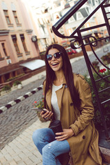 Fototapeta na wymiar Coffee break in her perfect city. Beautiful young woman in trench holding coffee cup and using her mobile phone with smile while sitting outdoors