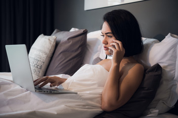 Fototapeta premium Beautiful woman lying in bed and making a phone call. Young girl using her laptop at home. Beautiful woman relaxing at home.