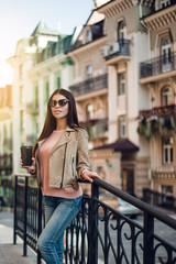 Beautiful lovely day. Full length of beautiful young woman in sunglasses holding coffee cup and looking at camera with smile while standing outdoors