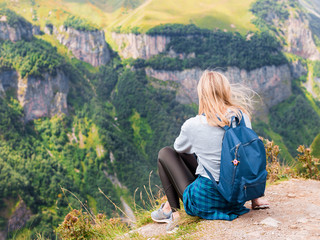 A girl sits on the edge of the cliff and looking at the valley. Woman enjoying the forest view from the top of the mountain in summer. The traveler girl at the peak of mountain.