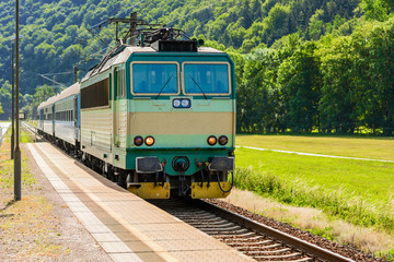 Obraz premium A green electric locomotive passing the Czech countryside. A train running through the green valley. Rail transport in the Czech Republic. The train arriving on a rural platform