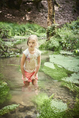 Fototapeta premium Child cute blond girl playing in the creek. Girl walking in forest stream and exploring nature. Summer children fun. Children summer adventure
