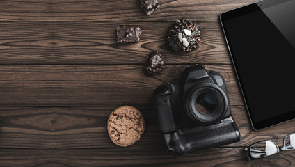 Top view travel concept with modern camera, digital tablet, eyeglasses and biscuits. Outfit of traveler on brown background with copy space