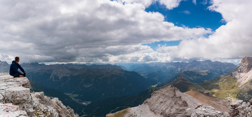 male hiker sitting on a mountain peak ledge in the Dolomites and looking at the amazing view of the surrounding landscape with Langkofel and Marmolada peaks