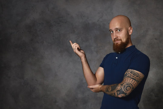 Attractive stylish European male with bushy beard, bald head and tattoo posing isolated in studio, pointing index finger, indicating copy space on gray wall for your advertisement or information