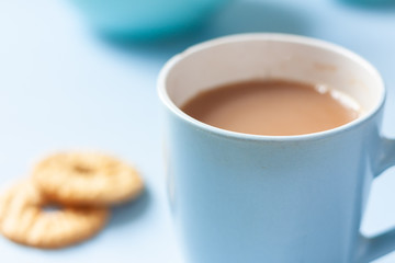 A blue mug of tea and biscuits with tea pot and accessories on a pale blue background