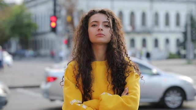 Confident strict cute curly teen girl portrait with arm crossed in yellow clothes, urban city background