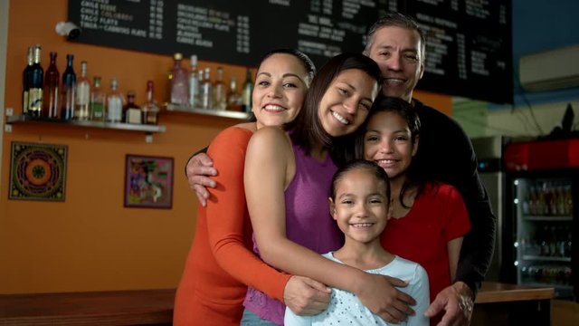 A beautiful hispanic family gives each other a group hug while looking into the camera in their family Mexican food restaurant.