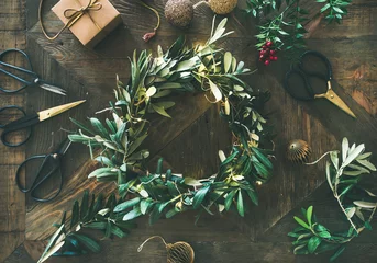 Fotobehang Getting ready for Christmas. Flat-lay of green olive branch festive wreath, Christmas tree decoration toys, gift box and scissors over rustic wood table background, top view, horizontal composition © sonyakamoz