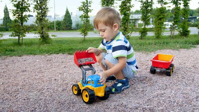 4k video of cute toddler boy playing on playground at park. Boy loading gravel with toy loader in trailer
