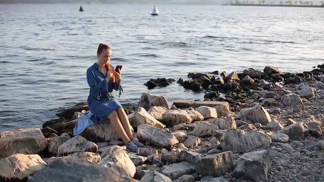 young beautiful Caucasian woman with red hair sitting on a large stone by the sea with a phone in her hand and communicate in social networks.