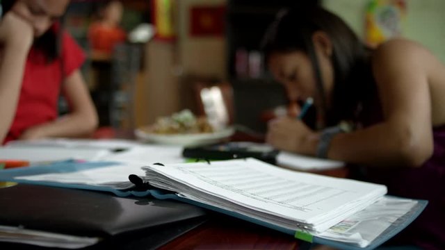 Close up shot of three young girls completing their homework, while their beautiful mother works the register in the background of their family owned restaurant