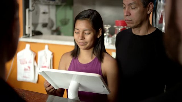 Supportive Hispanic father watches cute teen daughter sweetly take a debit card from customers, swipe it through the touch screen register, and turn the screen around for a signature while practicing the register at their family owned restaurant