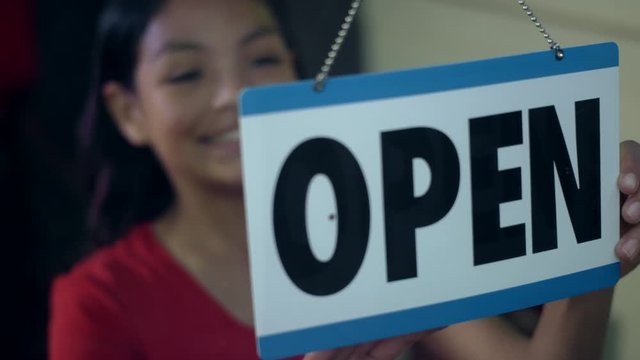 A young girl of hispanic descent turns a closed sign to open in the doorway window of her family's business