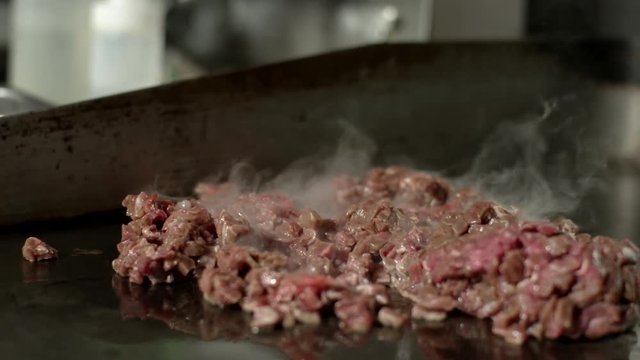 Close up of raw carne asada cooking on a flat grill. A chef tops it with onions and salt in a Mexican food restaurant kitchen