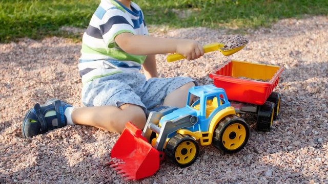Closeup 4k video of little toddler boy loading gravel in toy trailer with plastic shovel