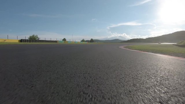 POV: Racing behind a red sports car speeding around the circuit on a sunny day. Following a bright colored supercar braking hard before a tight turn on asphalt racetrack. Adrenaline filled track day.