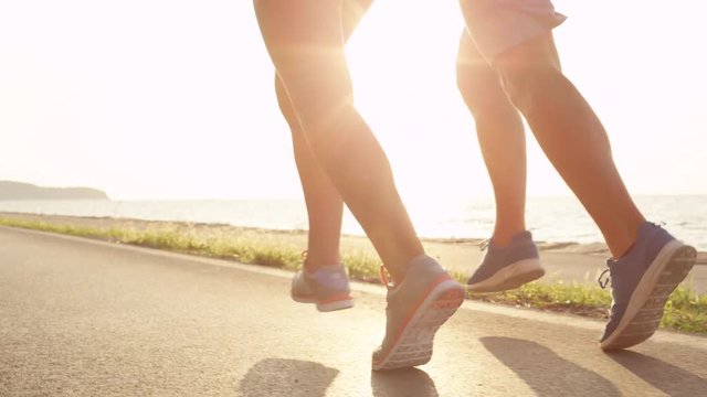 LENS FLARE, LOW ANGLE: Unknown young woman and man with athletic legs jogging in the sun down the scenic coastal pedestrian trail. Unrecognizable couple training together by the sea on a sunny day.