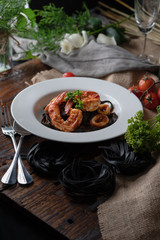 Black fettuccine spaghetti pasta with shrimp and squid topping with green parsley in white dish on woodden table with white wine and flowers, selective focus