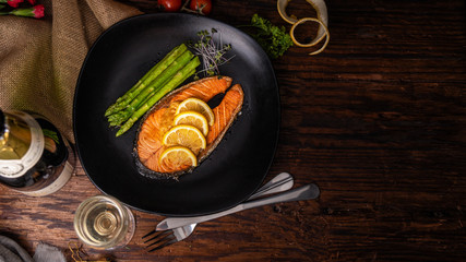salmon steak with green asparagus and topping with sliced yellow lemon on black plate on woodden...