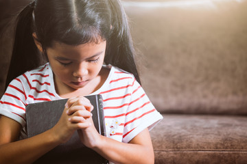 Cute asian little child girl closed her eyes and folded her hand in prayer on a Holy Bible for faith concept