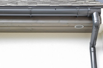 Grey plastic rain gutter with drain downspout pipe installed on asphalt shingles roof wooden eaves with round ventilation grille with copy space for text on white facade. Home improvement concept.