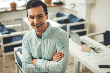 Ready to work. Delighted nice man smiling to you while standing cross handed in the office