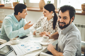 Our success. Delighted nice man smiling to you while working together with his colleagues