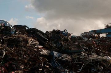 Pile of scrap metal at a junk yard with a metal structure on the side and a cloudy sky in the background