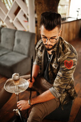 Stylish beard arabian man in glasses and military jacket smoking hookah at street bar. Arab model having rest.